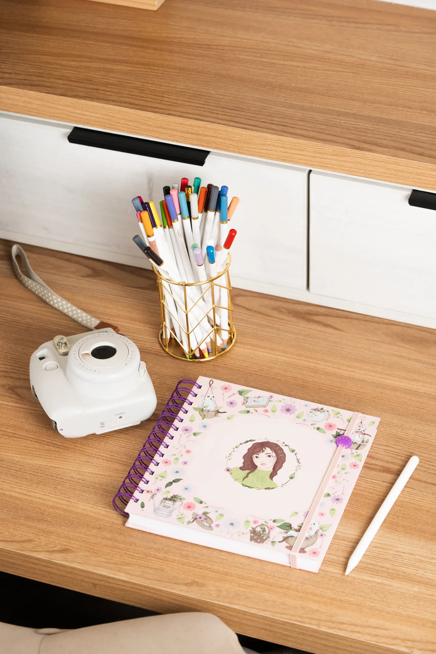 Cuaderno ilustrado OLFFY con lápices de colores y cámara instantánea sobre un escritorio de madera.